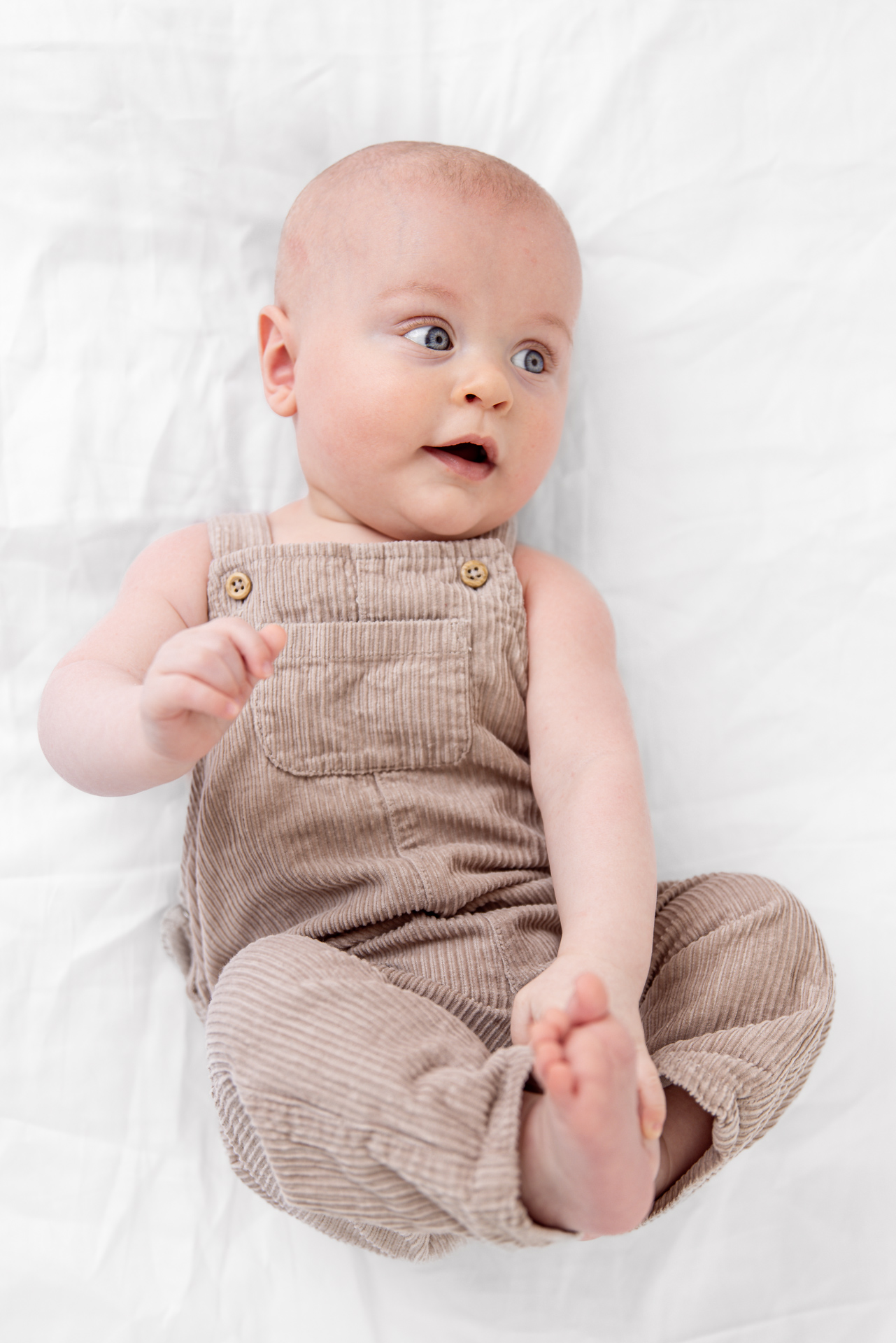Baby på 6 måneder ikledd snekkerbukse i cordfløyen, ligger på rygg, holder fast i beina og ser ut i rommet